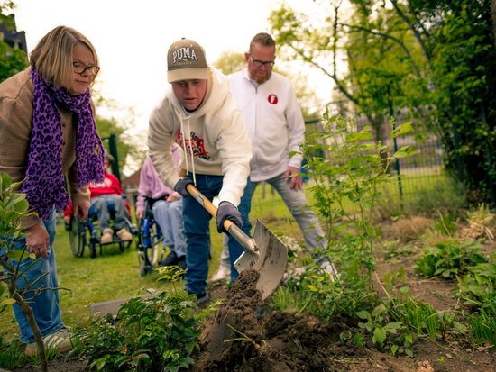 Drei Personen arbeiten in einem Garten. Ein Mann in der Mitte gräbt mit einer Schaufel Erde um, während eine Frau mit lila Schal links und ein Mann rechts zuschauen. Im Hintergrund sitzen zwei weitere Personen in Rollstühlen.