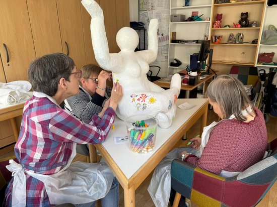 Drei Frauen sitzen an einem Tisch und bemalen gemeinsam eine große, weiße Skulptur einer Frau mit erhobenen Armen. Sie nutzen bunte Filzstifte aus einem Behälter, um kindliche Zeichnungen auf die Oberfläche der Figur zu bringen.