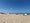 Ein sonniger Tag am Strand unter einem klaren blauen Himmel. Menschen entspannen im Sand, einige liegen zum Sonnenbaden. Eine gelbe Flagge auf einem Mast signalisiert die Badebedingungen, und das Meer erstreckt sich im Hintergrund.