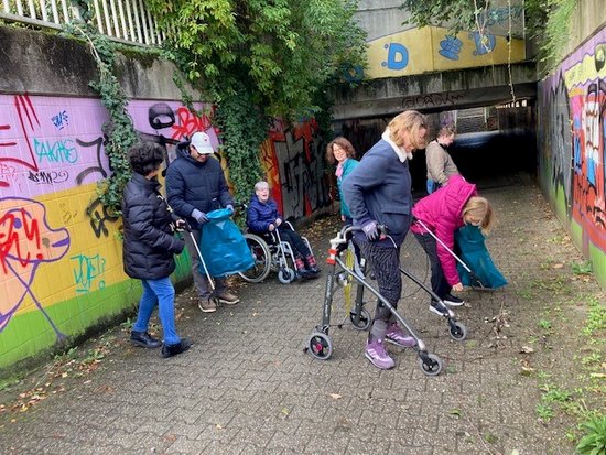 Menschen unterschiedlichen Alters, darunter ein Kind im Rollstuhl und eine Seniorin mit Rollator, räumen Müll in einer graffitibeschmierten Unterführung auf.