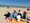 Eine Gruppe von fünf Menschen sitzt im Kreis auf Handtüchern am Strand und unterhält sich. Im Hintergrund sind das Meer und weitere Strandbesucher unter einem klaren blauen Himmel zu sehen.