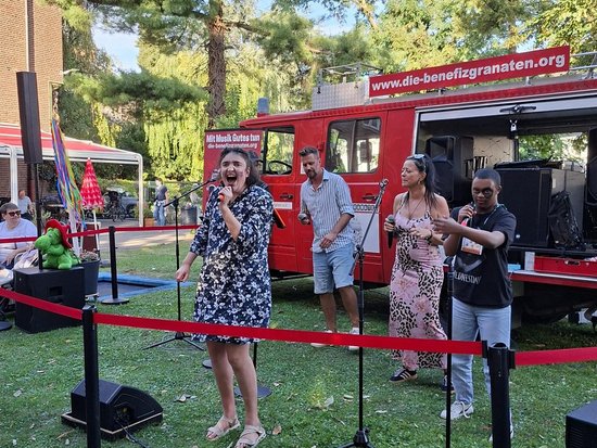 Eine Sängerin steht im Vordergrund und singt in ein Mikrofon, begleitet von Musikern auf einer Wiese. Im Hintergrund ist ein roter Truck mit der Webadresse www.die-benefizgranaten.org zu sehen.