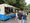 Ein blauer und weißer Food Truck steht auf einem Weg im Freien. Mehrere Menschen warten in einer Schlange vor dem geöffneten Schalter. Im Inneren des Fahrzeugs sind Angestellte beim Arbeiten zu sehen. Im Hintergrund sind Bäume und weitere Besucher erkennbar.