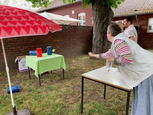 Eine Frau wirft bei einem Wurfspiel im Garten. Vor ihr steht ein Tisch mit Bechern, daneben ein roter Schirm mit Herz-Muster. Zwei Männer beobachten das Spiel im Hintergrund.