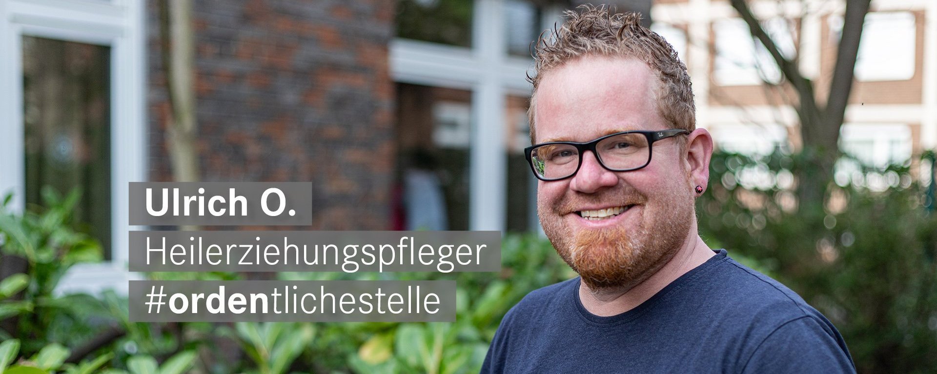 Heilerziehungspfeger mit orangen Haaren und Brille laechelt freundlich in die Kamera