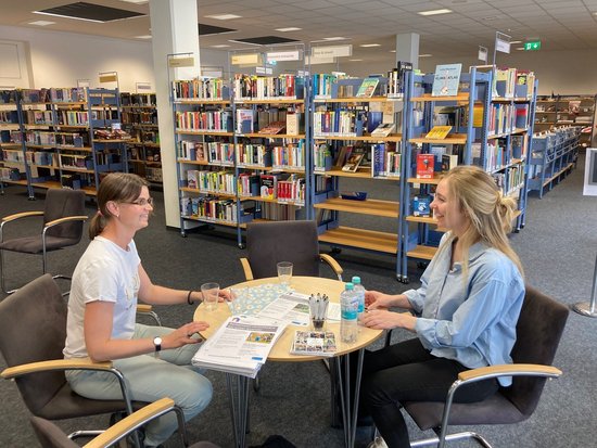 Zwei junge Frauen unterhalten sich an einem runden Tisch in einer Bibliothek. Im Hintergrund sind zahlreiche Bücherregale zu erkennen. Auf dem Tisch liegen Dokumente und Getränke.