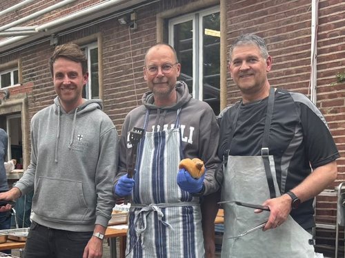 Drei Männer stehen lächelnd hinter einem langen Grill, auf dem Würstchen und Burger zubereitet werden. Der Mann in der Mitte hält ein Brötchen, der Mann rechts Grillzangen. Im Hintergrund sind ein Backsteingebäude und weitere Personen zu sehen.