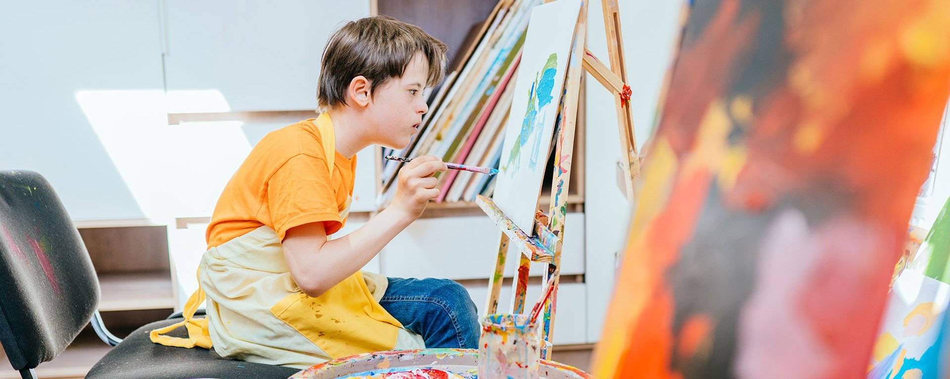 Kreativ: Ein Junge sitzt an einer Staffelei und malt mit Acrylfarben konzentriert ein Bild