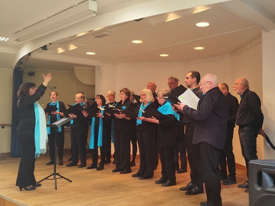 Ein gemischter Chor steht auf einer Holzbühne und singt. Die Sänger tragen schwarze Kleidung, die Frauen zusätzlich helle blaue Schals. Eine Dirigentin links leitet die Aufführung mit erhobenem Arm. Alle halten schwarze Notenordner. Blaue Vorhänge rahmen die Bühne ein.
