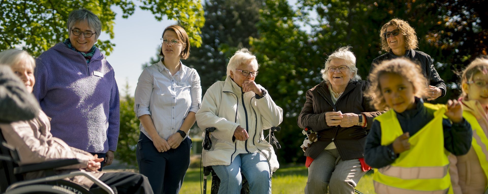 Mitarbeitende, Senioren und Kinder stehen auf einer Wiese und unterhalten sich.