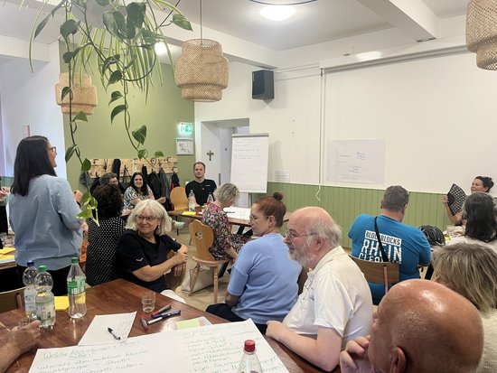 Eine Gruppe von Menschen sitzt in einem hellen Raum an Tischen und nimmt an einer Diskussion oder einem Workshop teil. Eine Frau steht links und spricht zu den anderen, während auf den Tischen Notizzettel und Stifte liegen.