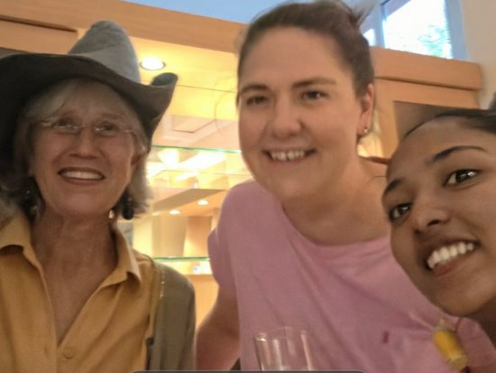 Vier lächelnde Frauen posieren eng beieinander für ein Selfie. Von links nach rechts: eine Frau mit Brille und rotem Schal, eine ältere Dame in einem gelben Hemd mit grauem Hut, eine Frau in Rosa und eine Frau mit großen gelben Ohrringen.