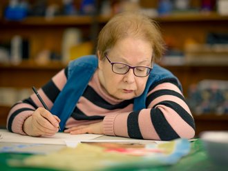 Eine ältere Dame mit Brille und gestreiftem Pullover schreibt konzentriert mit einem Stift auf Papier. Sie trägt eine blaue Weste über dem Pullover. Im Hintergrund sind unscharfe Bücherregale sichtbar.