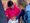 Eine Person in einer rosa Jacke sitzt auf einem roten Stuhl und hält ein braunes Kaninchen auf einem weißen Tuch auf dem Schoß. Eine zweite Person in einer blauen Jacke streichelt das Tier sanft. Im Hintergrund sind weitere rote Plastikstühle und eine Ziegelmauer sichtbar.