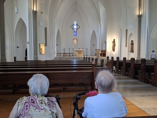 Zwei ältere Frauen sitzen auf einer Kirchenbank und schauen zum Altar. Die Kirche hat hohe weiße Wände und Gewölbedecken. Eine Frau hat einen Rollator neben sich. Im Hintergrund hängt ein leuchtendes Kreuz.