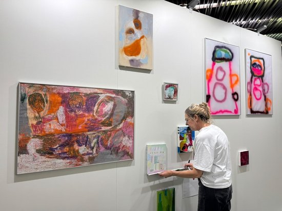 Eine Frau in weißem T-Shirt steht in einer Kunstgalerie vor einer weißen Wand und betrachtet ein kleines abstraktes Gemälde. Um sie herum hängen weitere bunte, expressionistische Werke unterschiedlicher Größe, darunter ein großes horizontales Bild links und zwei vertikale Bilder rechts.