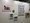 Eine weiße Galeriewand präsentiert diverse Kunstwerke. Links hängt ein abstraktes Bild mit bunten Formen, daneben ein kleines farbiges Objekt. Rechts befindet sich ein großes Poster mit vielen kleinen Motiven. Darunter sind zwei kleinere Bilder in Blautönen zu sehen.