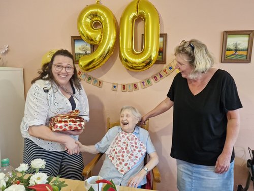 Eine ältere Frau feiert ihren 90. Geburtstag. Sie sitzt lachend zwischen zwei Frauen. Eine reicht ihr ein Geschenk, die andere steht lächelnd daneben. Goldene Ballons mit der Zahl 90 und eine Girlande schmücken den Hintergrund.