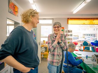 Zwei Frauen stehen in einem hellen Atelier und unterhalten sich. Die Frau im karierten Hemd klatscht in die Hände, während die andere lächelt. Im Hintergrund sitzt eine weitere Person an einem Tisch und arbeitet.