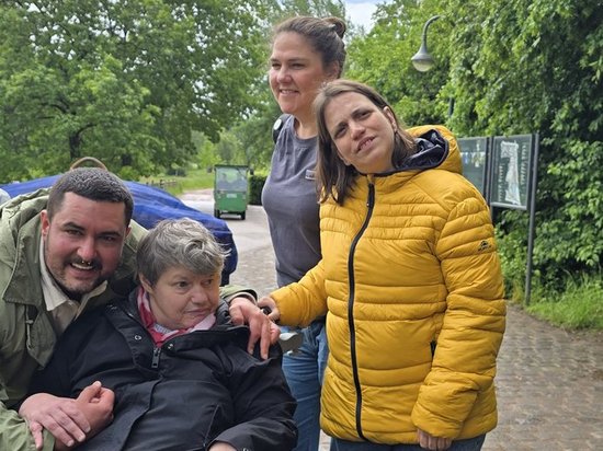 Vier fröhliche Personen posieren im Park. Eine ältere Frau sitzt im Rollstuhl, ein Mann lehnt sich von links zu ihr, und zwei Frauen stehen rechts daneben. Alle lächeln in die Kamera vor einem Hintergrund aus Bäumen und blauem Himmel.