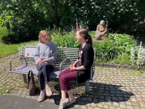 Zwei junge Frauen sitzen sich auf einer Parkbank gegenüber und unterhalten sich. Im Hintergrund ist ein grüner Gartenbereich mit Blumen und einer sitzenden Skulptur zu sehen.