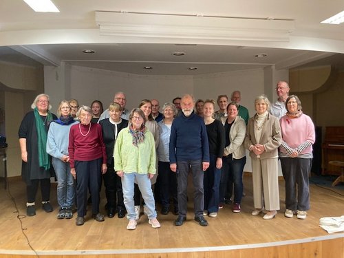 Ein Gruppenfoto von etwa 20 Personen, überwiegend älteren Erwachsenen, die auf einer Holzbühne in einem hellen Raum stehen. Sie tragen alltägliche Freizeitkleidung und blicken freundlich in die Kamera.