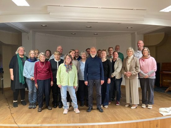 Ein Gruppenfoto von etwa 20 Personen, überwiegend älteren Erwachsenen, die auf einer Holzbühne in einem hellen Raum stehen. Sie tragen alltägliche Freizeitkleidung und blicken freundlich in die Kamera.