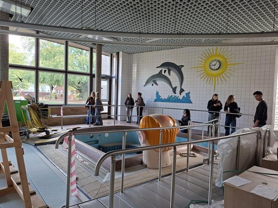 Ein Hallenbad während der Renovierung. Im Vordergrund steht eine große Kopf-Skulptur im leeren Becken. An der weißen Fliesenwand sind zwei springende Delfine und eine große gelbe Sonnen-Uhr zu sehen. Eine Gruppe von Menschen betrachtet den Raum, links sind Baugeräte und Fenster sichtbar.