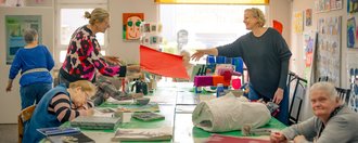 In einem hellen Raum arbeiten mehrere ältere Menschen gemeinsam an Kunstprojekten. Sie sitzen oder stehen um einen langen Tisch, der mit Papier und Materialien bedeckt ist. Zwei Frauen halten ein rotes Blatt Papier hoch. An den Wänden hängen bunte Gemälde.