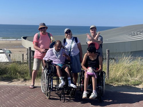 Eine Gruppe von fünf Menschen posiert an einem sonnigen Tag am Strand. Zwei Personen sitzen in Rollstühlen, drei stehen dahinter. Im Hintergrund sind das Meer und ein modernes Gebäude mit gewölbtem Dach zu sehen. Die Stimmung ist fröhlich.