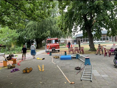 Ein gepflasterter Platz mit einem roten Feuerwehrwagen im Hintergrund. Im Vordergrund liegen verschiedene Gegenstände verstreut: eine Leiter, ein langer Schlauch, eine blaue Matte und Holz-Hindernisse. Links stehen zwei Frauen, rechts sitzen Zuschauer auf roten Stühlen.