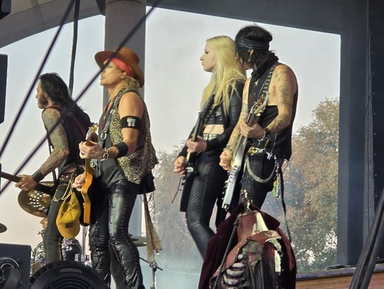 Vier Musiker einer Rockband stehen auf einer überdachten Außenbühne. Von links nach rechts: ein Gitarrist mit Resonatorgitarre, ein Sänger mit Cowboyhut und Leopardenweste, eine blonde Gitarristin in schwarzer Lederkleidung und ein Gitarrist mit Bandana und weißer E-Gitarre.